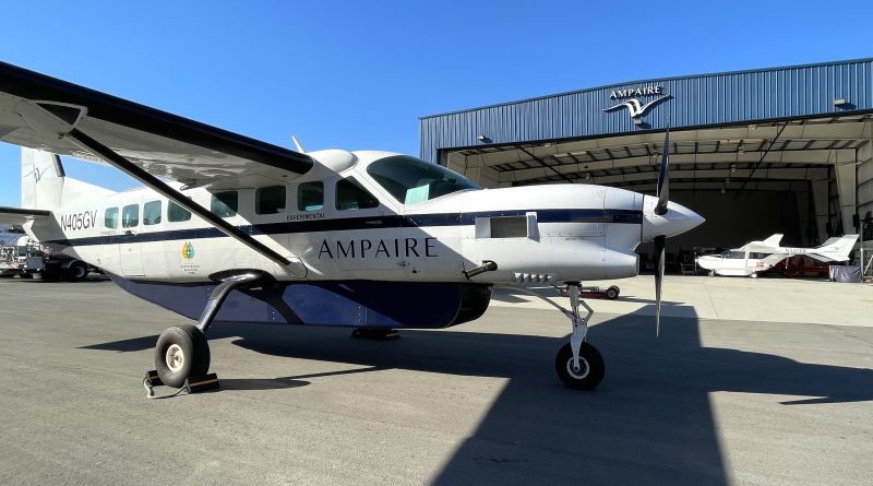 Small white and blue experimental propeller airplane on the tarmac in front of a blue AMPAIRE hangar.”
