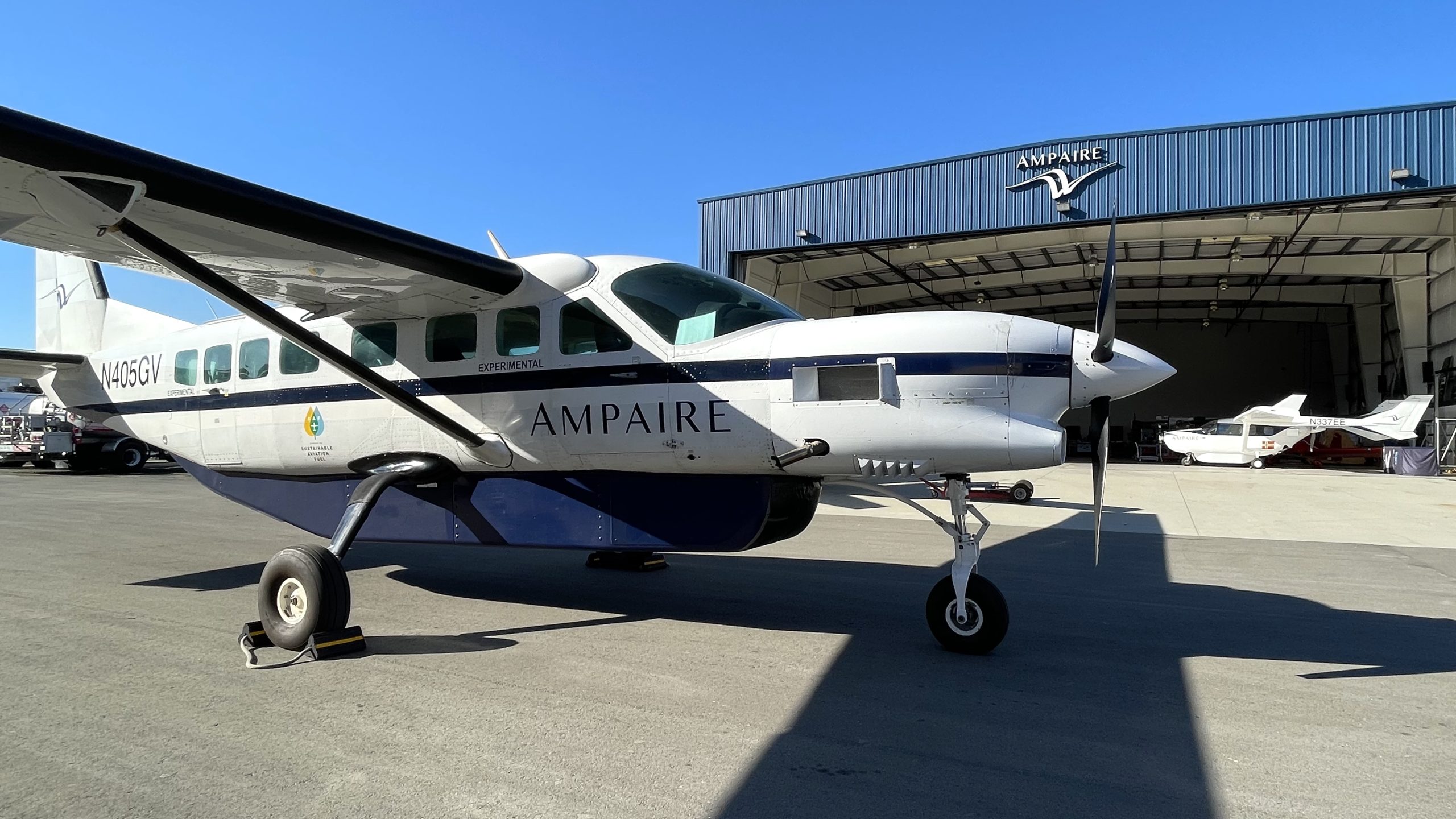 Small white and blue experimental propeller airplane on the tarmac in front of a blue AMPAIRE hangar.”