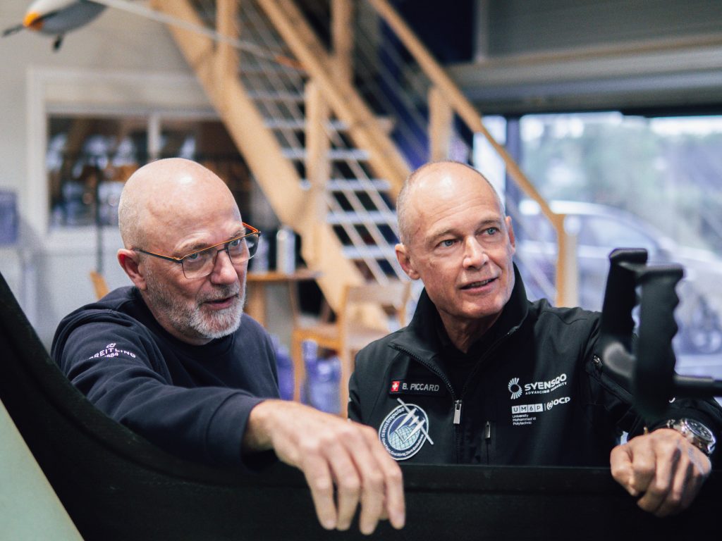 Two bald men in a workshop lean over a workstation, looking at a screen together, with stairs in the background.