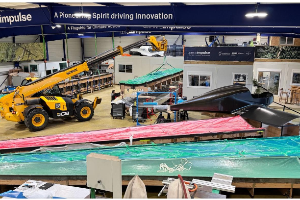 Industrial workshop with a yellow JCB telehandler lifting a component over a sleek black experimental aircraft,workbenches and banners in the background