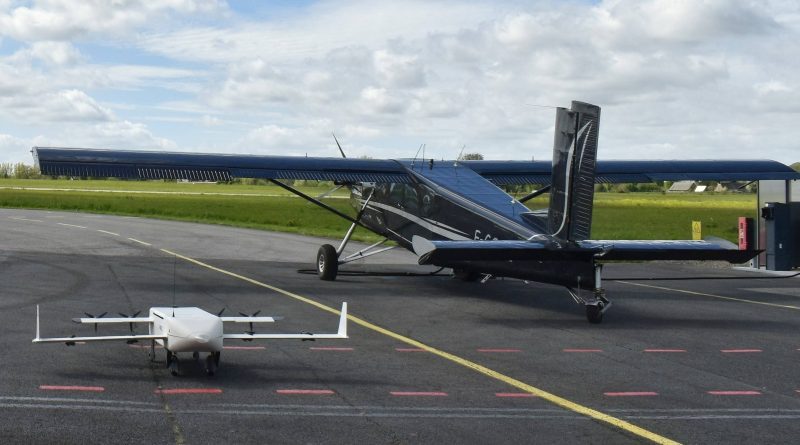Small white quadcopter drone sits on the tarmac in the foreground, with a blue single-engine airplane parked behind it on the apron.