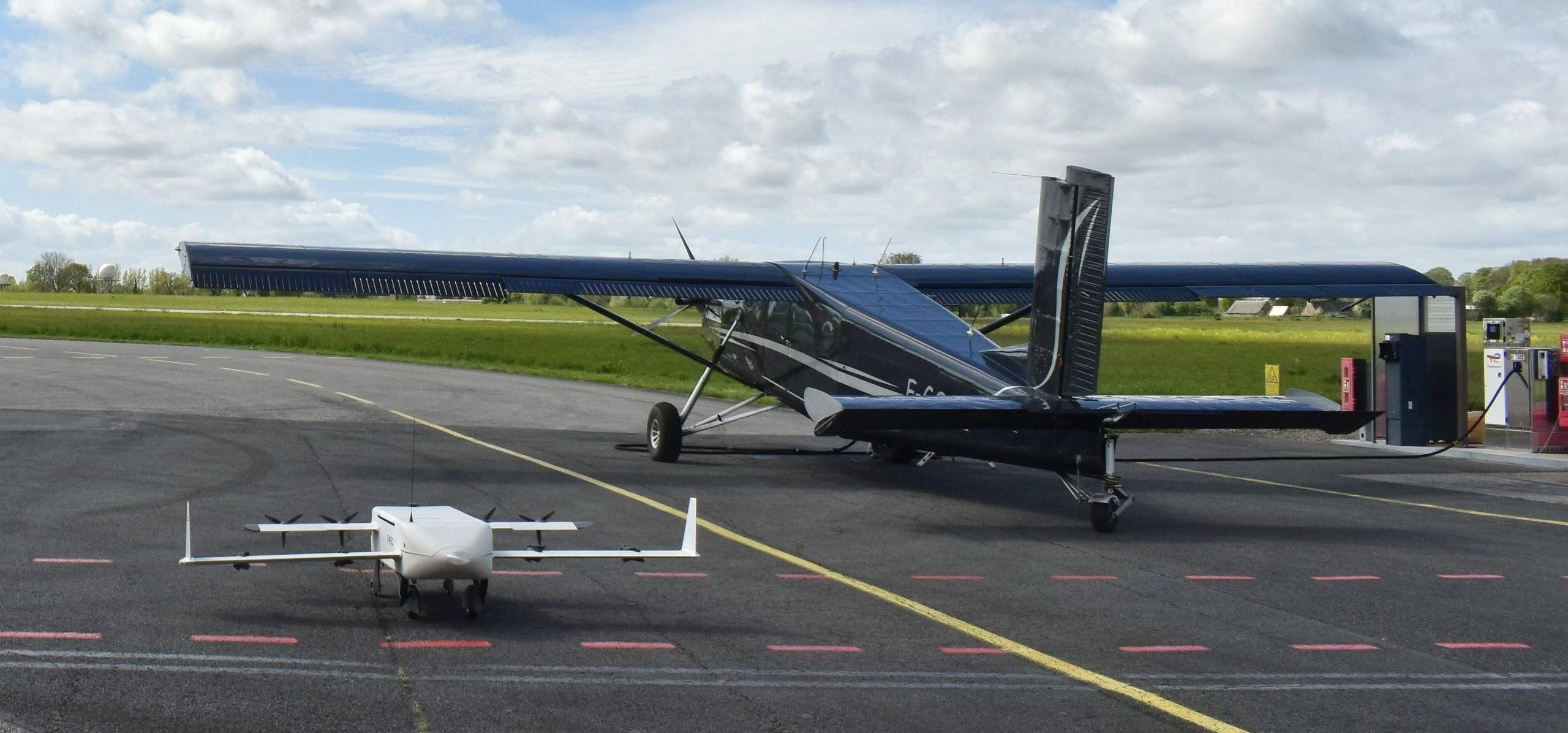 Small white quadcopter drone sits on the tarmac in the foreground, with a blue single-engine airplane parked behind it on the apron.