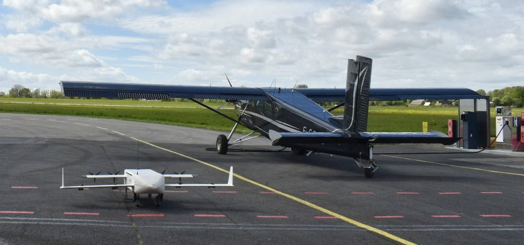 DSC_0764~2 3 - eVTOL Insights White small drone on a tarmac in the foreground with a dark blue ultralight airplane parked behind near fuel pumps.