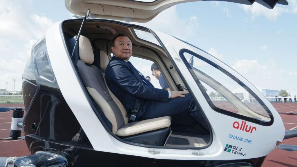 EH216-S 1 - eVTOL Insights Man in a dark suit sits in the open cockpit of a small, futuristic helicopter on a runway, nose with logos visible.