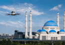 White mosque with blue domes and four minarets under a blue sky; a helicopter flies by.