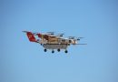 Large multi-rotor drone with a red tail and white body flying in a clear blue sky.