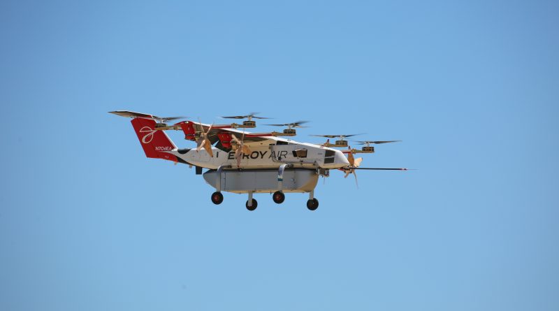 Large multi-rotor drone with a red tail and white body flying in a clear blue sky.