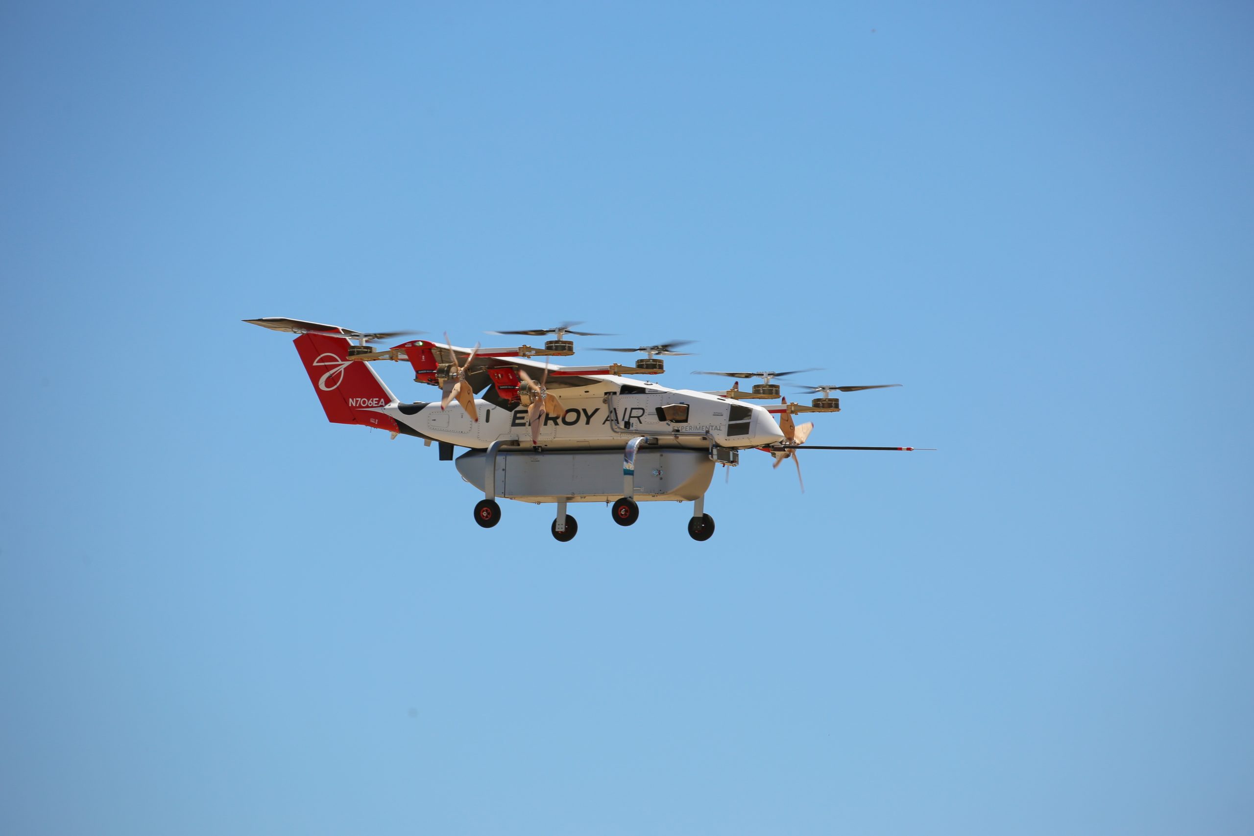 Large multi-rotor drone with a red tail and white body flying in a clear blue sky.