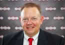 Portrait of a smiling man in a dark suit and red tie, standing in front of a branded backdrop with repeating logos.