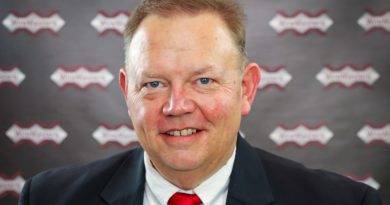 Portrait of a smiling man in a dark suit and red tie, standing in front of a branded backdrop with repeating logos.