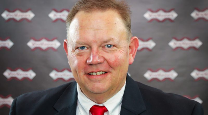 Portrait of a smiling man in a dark suit and red tie, standing in front of a branded backdrop with repeating logos.