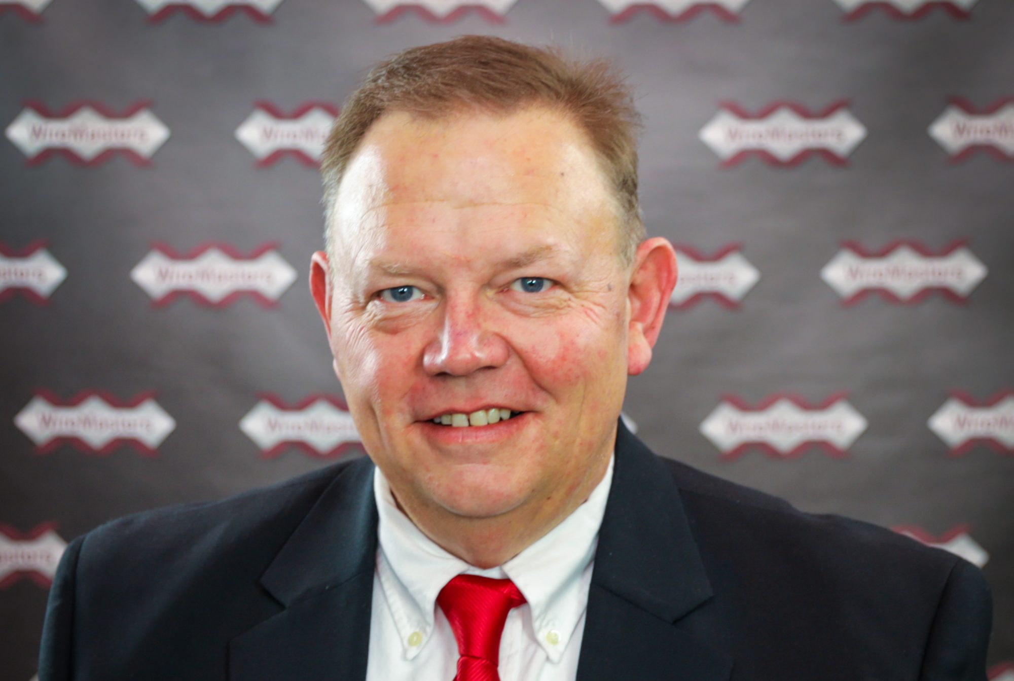 Portrait of a smiling man in a dark suit and red tie, standing in front of a branded backdrop with repeating logos.