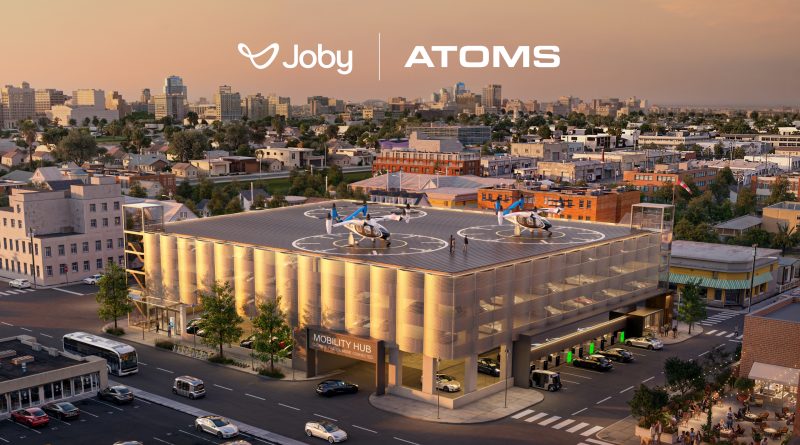 Aerial view of a city with two small helicopters on a rooftop heliport and a large Joby ATOMS banner across the sky above.