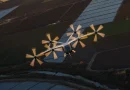 Six-propeller fixed-wing drone flying over farmland, sunlight glinting on the fuselage and wings.