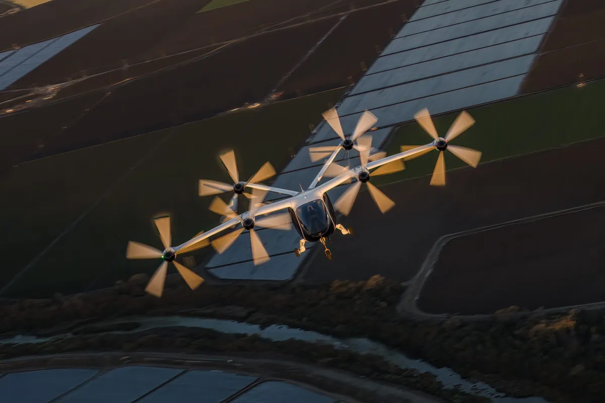 Six-propeller fixed-wing drone flying over farmland, sunlight glinting on the fuselage and wings.