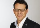 Portrait of a man with dark hair and rectangular glasses, wearing a navy blazer and white shirt, smiling.