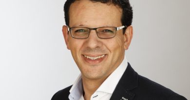 Portrait of a man with dark hair and rectangular glasses, wearing a navy blazer and white shirt, smiling.