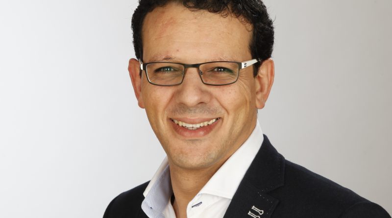 Portrait of a man with dark hair and rectangular glasses, wearing a navy blazer and white shirt, smiling.