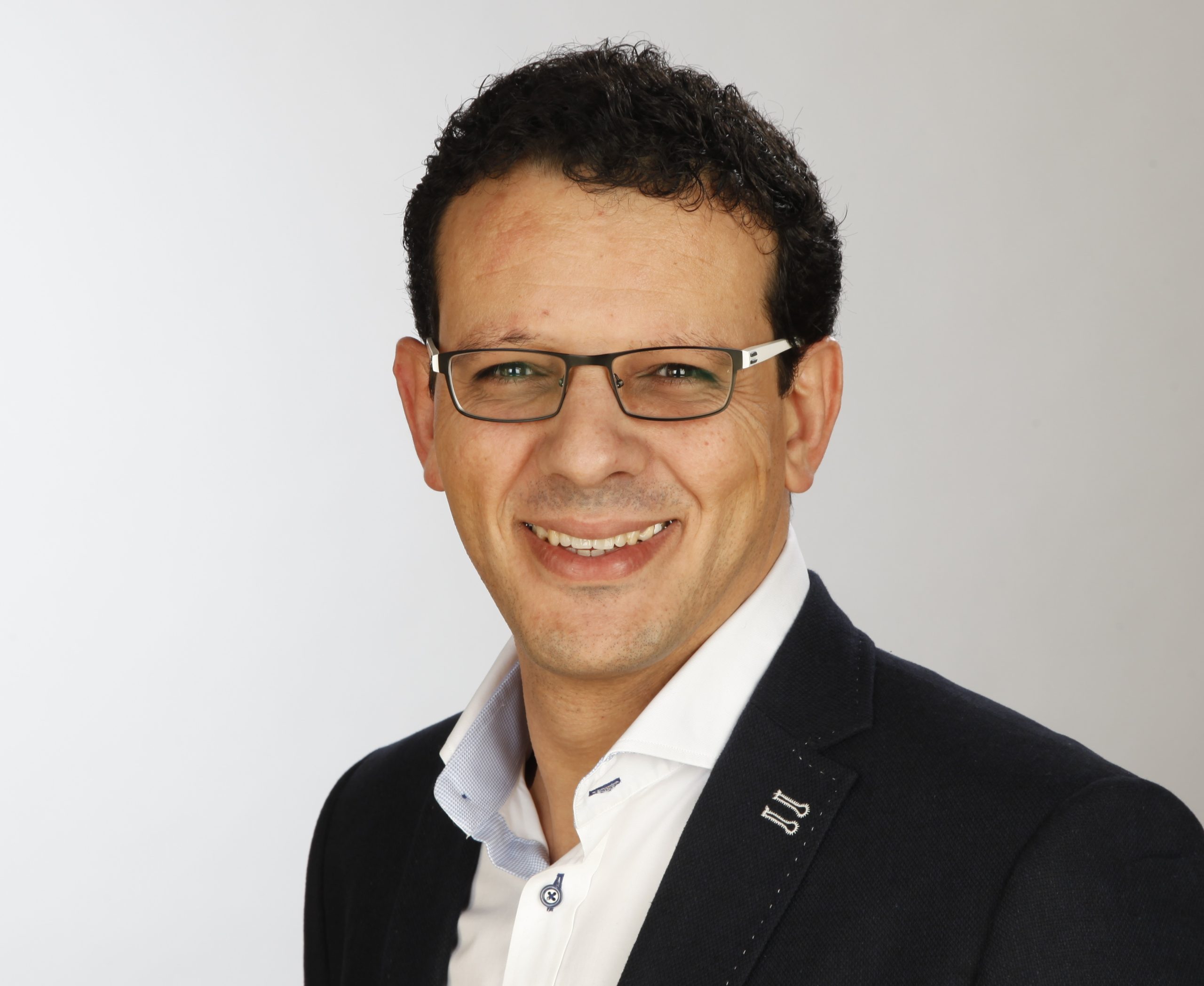 Portrait of a man with dark hair and rectangular glasses, wearing a navy blazer and white shirt, smiling.