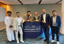 Six men pose behind a purple banner that reads 'Advanced Air Mobility Forum' in English and Arabic, at a conference retreat setting.