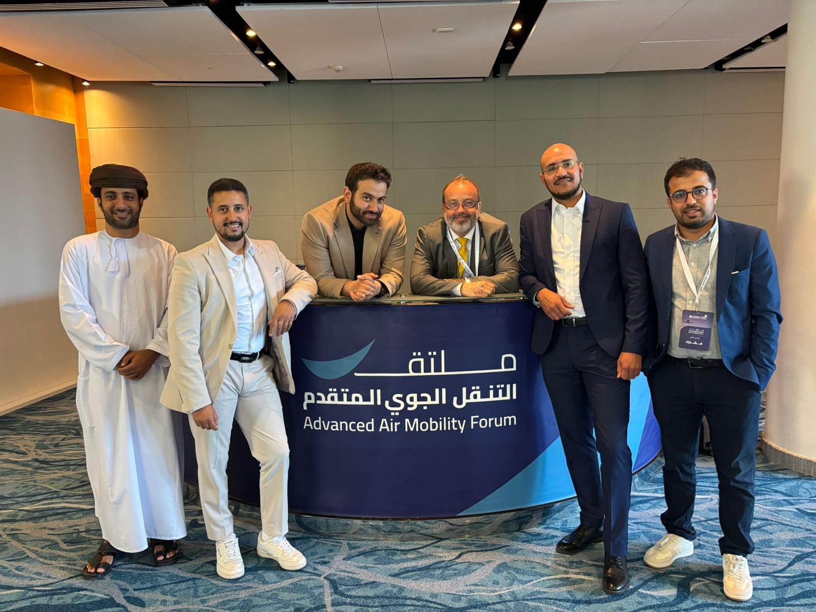 Six men pose behind a purple banner that reads 'Advanced Air Mobility Forum' in English and Arabic, at a conference retreat setting.