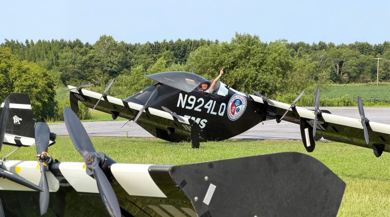 Person in a black aircraft cockpit raises one arm in a thumbs-up gesture beside a small propeller plane on a grassy field with trees in the background.