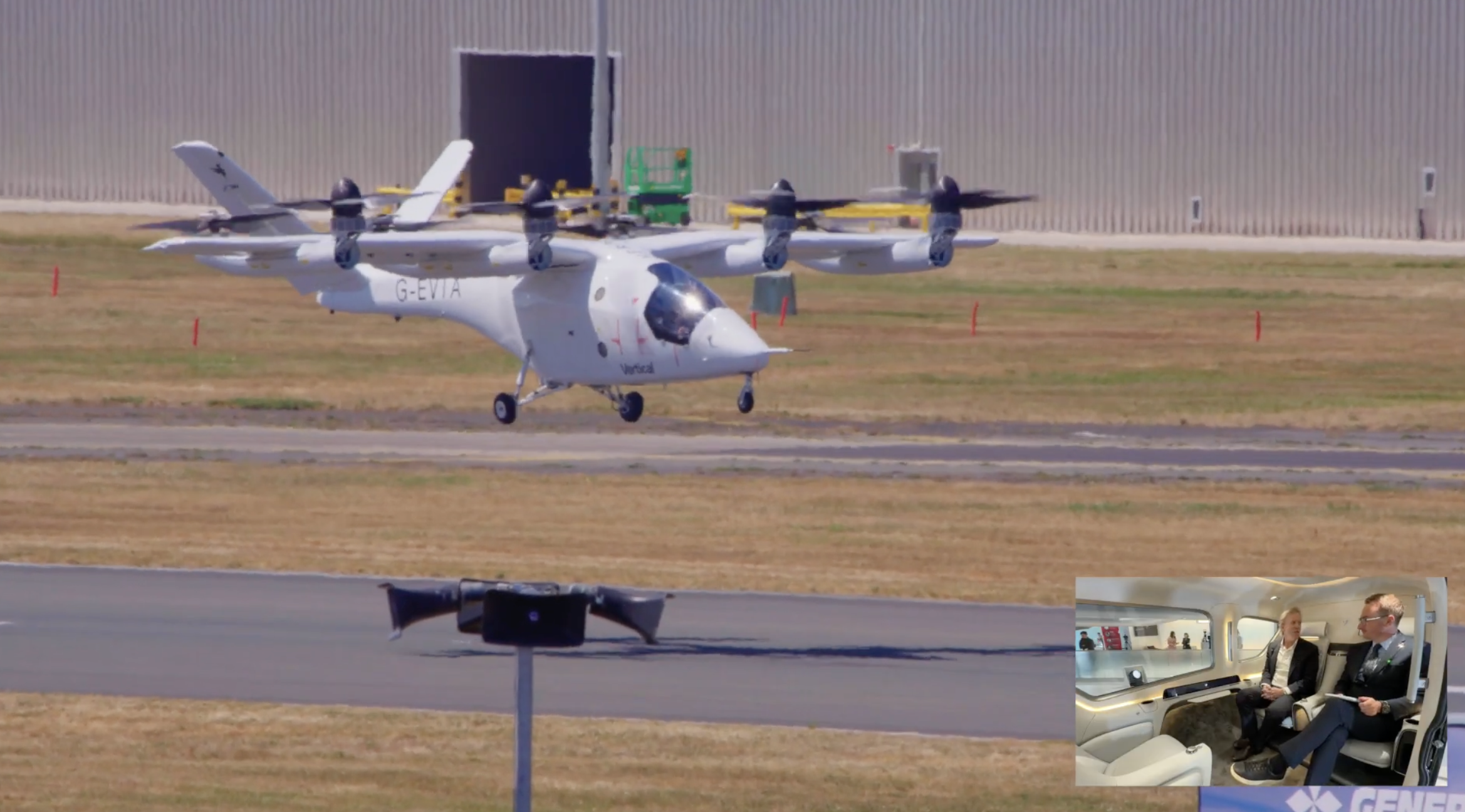 White tilt-rotor aircraft with four rotors taxiing on a runway; an inset photo in the lower-right shows two people seated inside a cabin. The aircraft bears registration G-EVIA and is near a hangar on a sunny day.