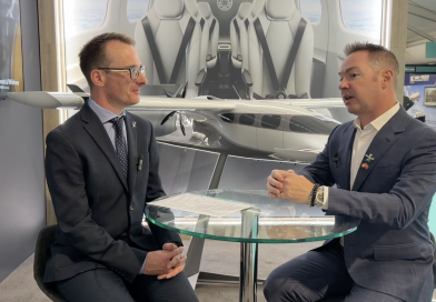 Two men in suits sit across a glass table, speaking in a showroom with a large airplane model behind them.