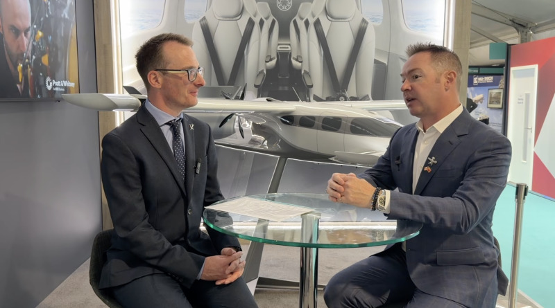 Two men in suits sit across a glass table, speaking in a showroom with a large airplane model behind them.