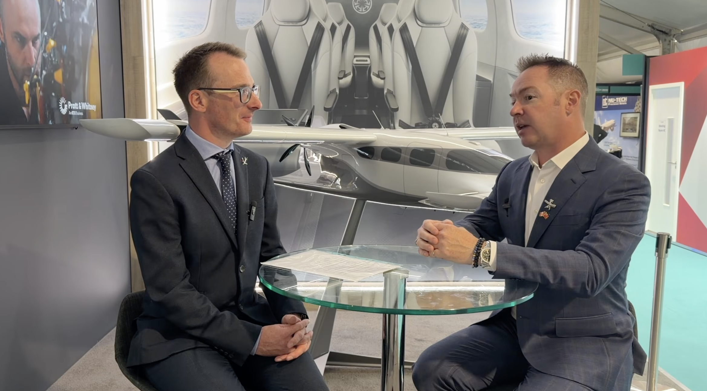 Two men in suits sit across a glass table, speaking in a showroom with a large airplane model behind them.