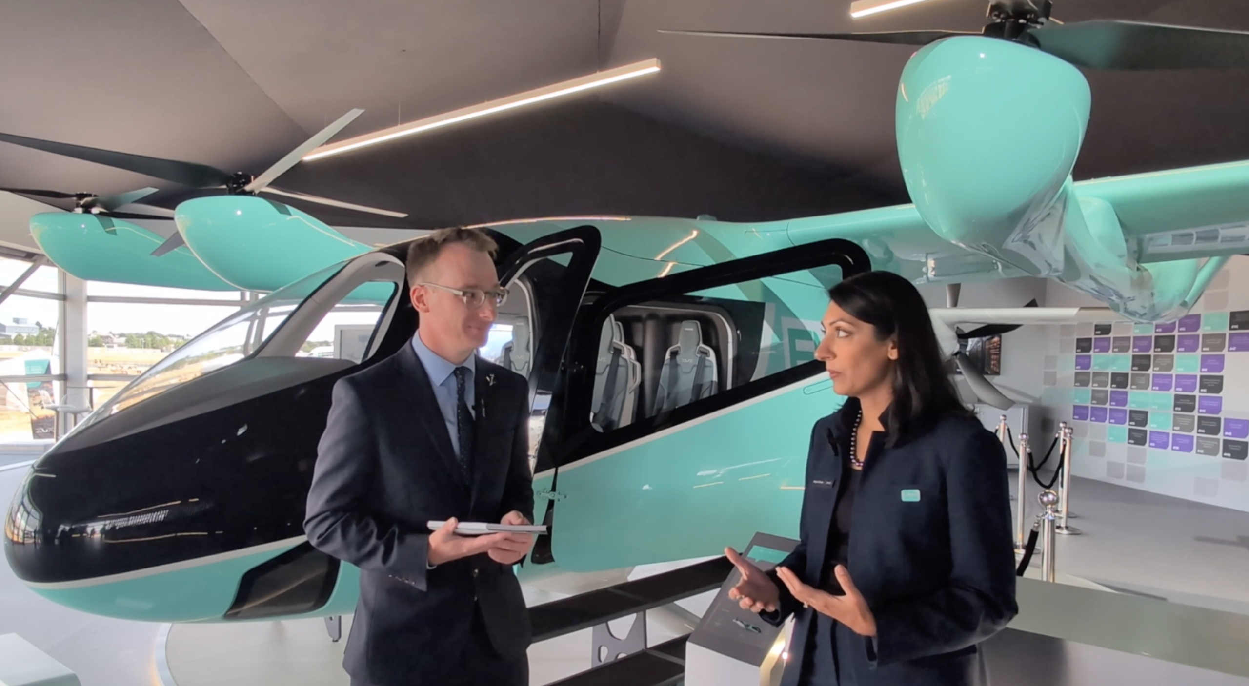 Two professionals in business attire stand beside a teal helicopter inside a showroom, talking.