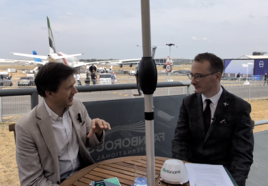 Two men in suits chat at an outdoor interview table on an airfield with airplanes in the background.Change is in the air banner visible behind them.