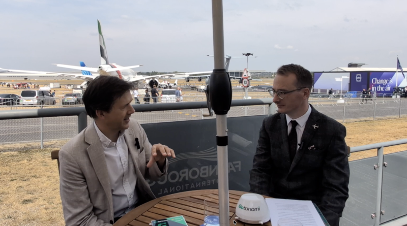 Two men in suits chat at an outdoor interview table on an airfield with airplanes in the background.Change is in the air banner visible behind them.