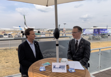 Two men in business attire seated at a round wooden table under a canopy at an outdoor aviation event, with parked airplanes in the background.