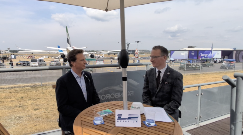 Two men in business attire seated at a round wooden table under a canopy at an outdoor aviation event, with parked airplanes in the background.
