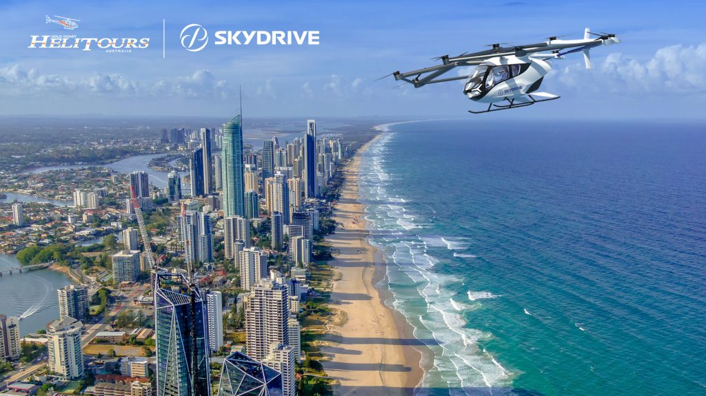 Aerial view of a coastal city skyline with high-rise buildings along a long sandy beach, and a white helicopter flying overhead with SkyDrive branding.