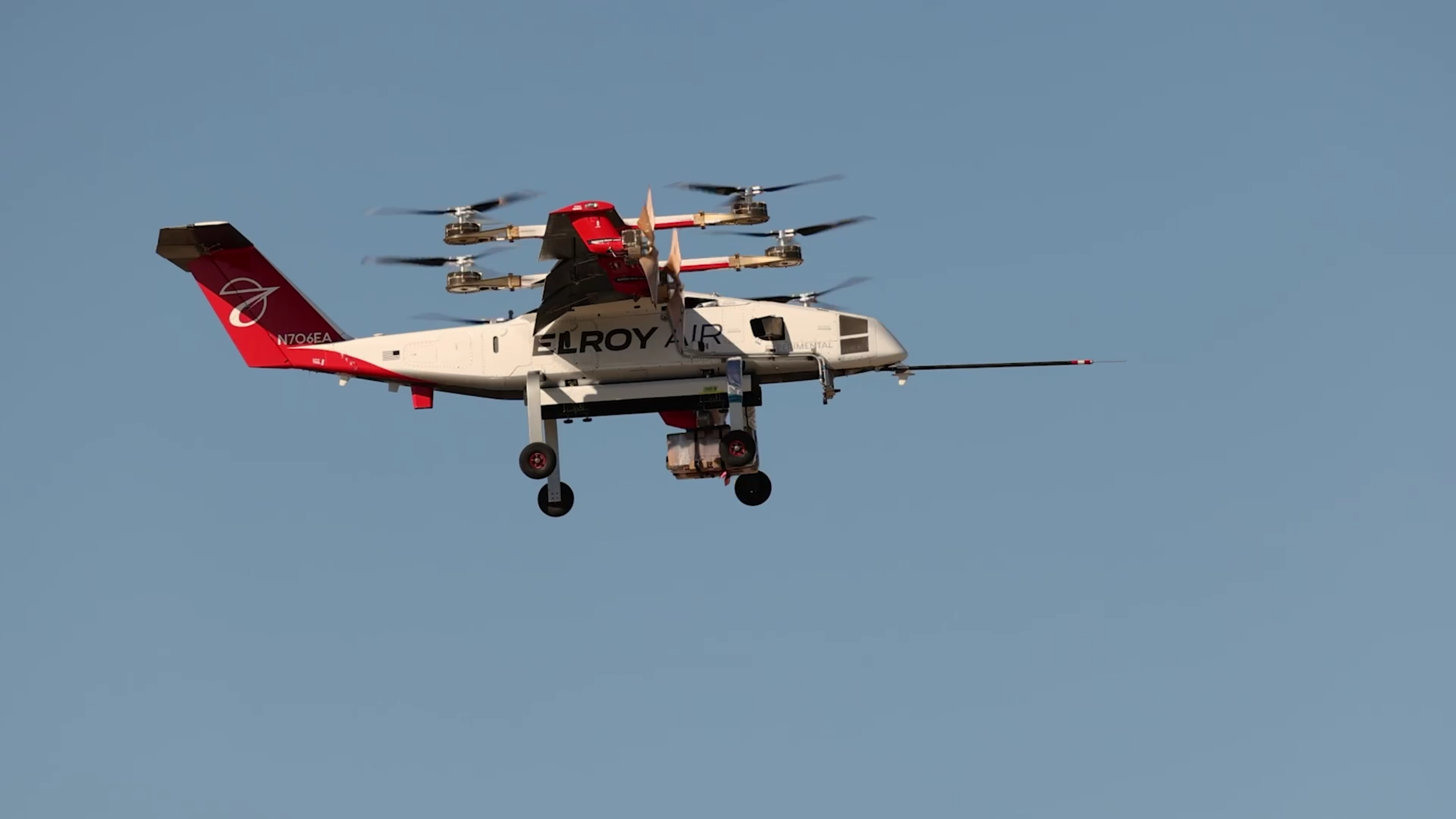 Elroy Air cargo drone with six rotor arms in midflight over a blue sky, carrying a payload beneath the fuselage.