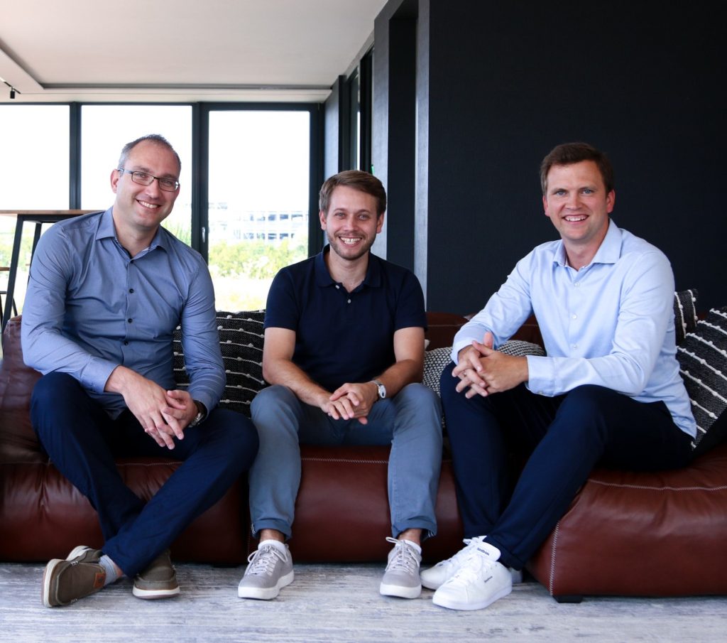 Three men sit closely on a brown leather sofa in a bright modern office, smiling at the camera.