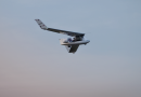 Small experimental aircraft flying in clear blue sky, cockpit visible with two occupants and wings angled upward