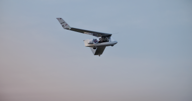 Small experimental aircraft flying in clear blue sky, cockpit visible with two occupants and wings angled upward