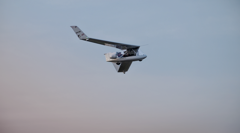 Small experimental aircraft flying in clear blue sky, cockpit visible with two occupants and wings angled upward