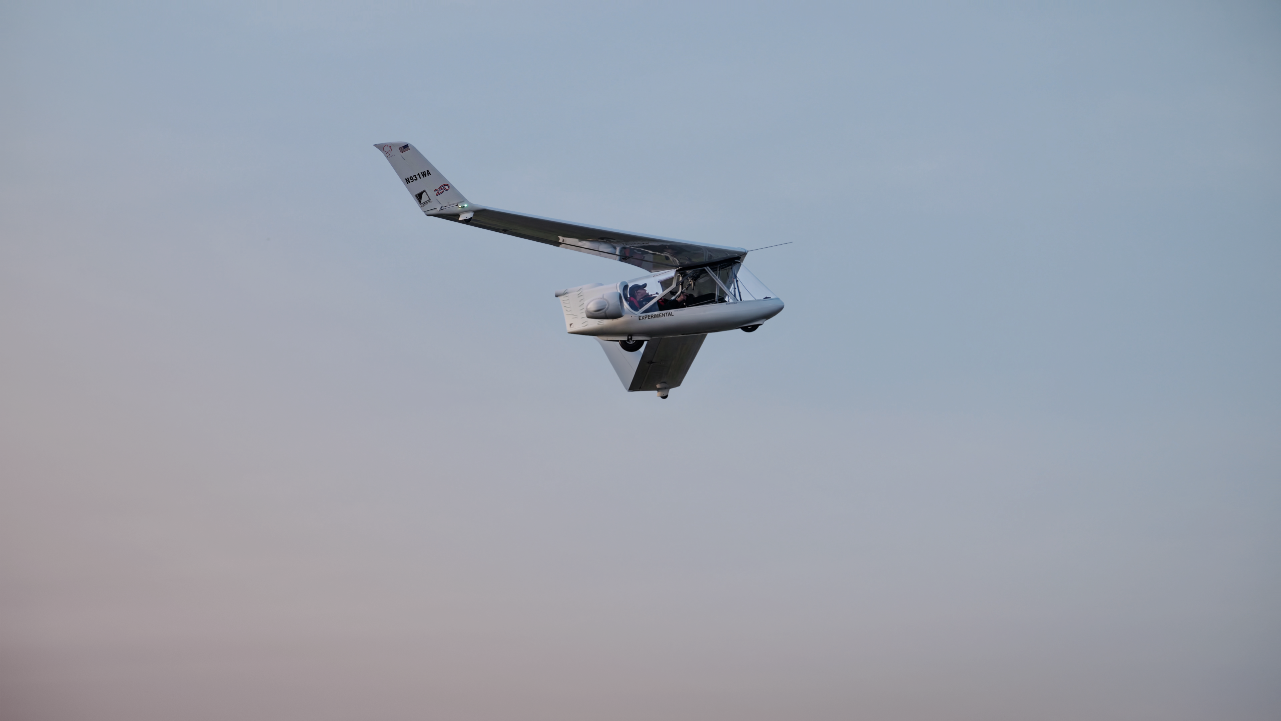 Small experimental aircraft flying in clear blue sky, cockpit visible with two occupants and wings angled upward