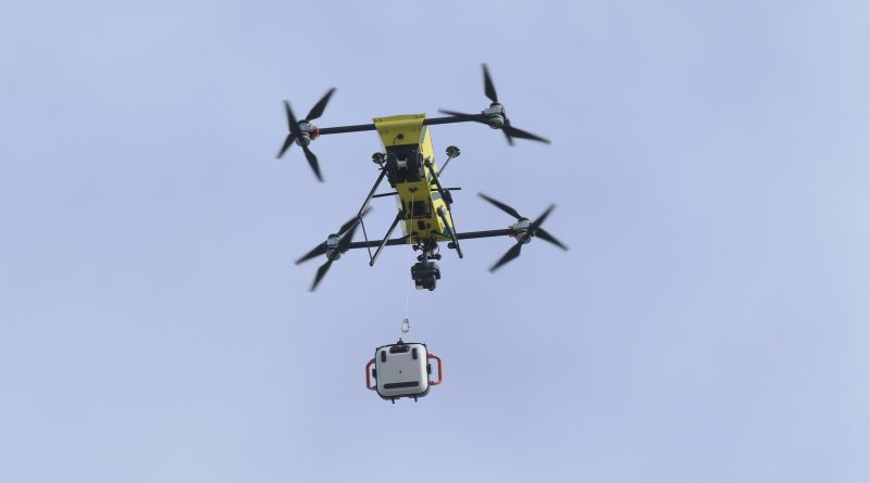 Yellow multi-rotor drone carrying a white container suspended beneath it against a clear blue sky.