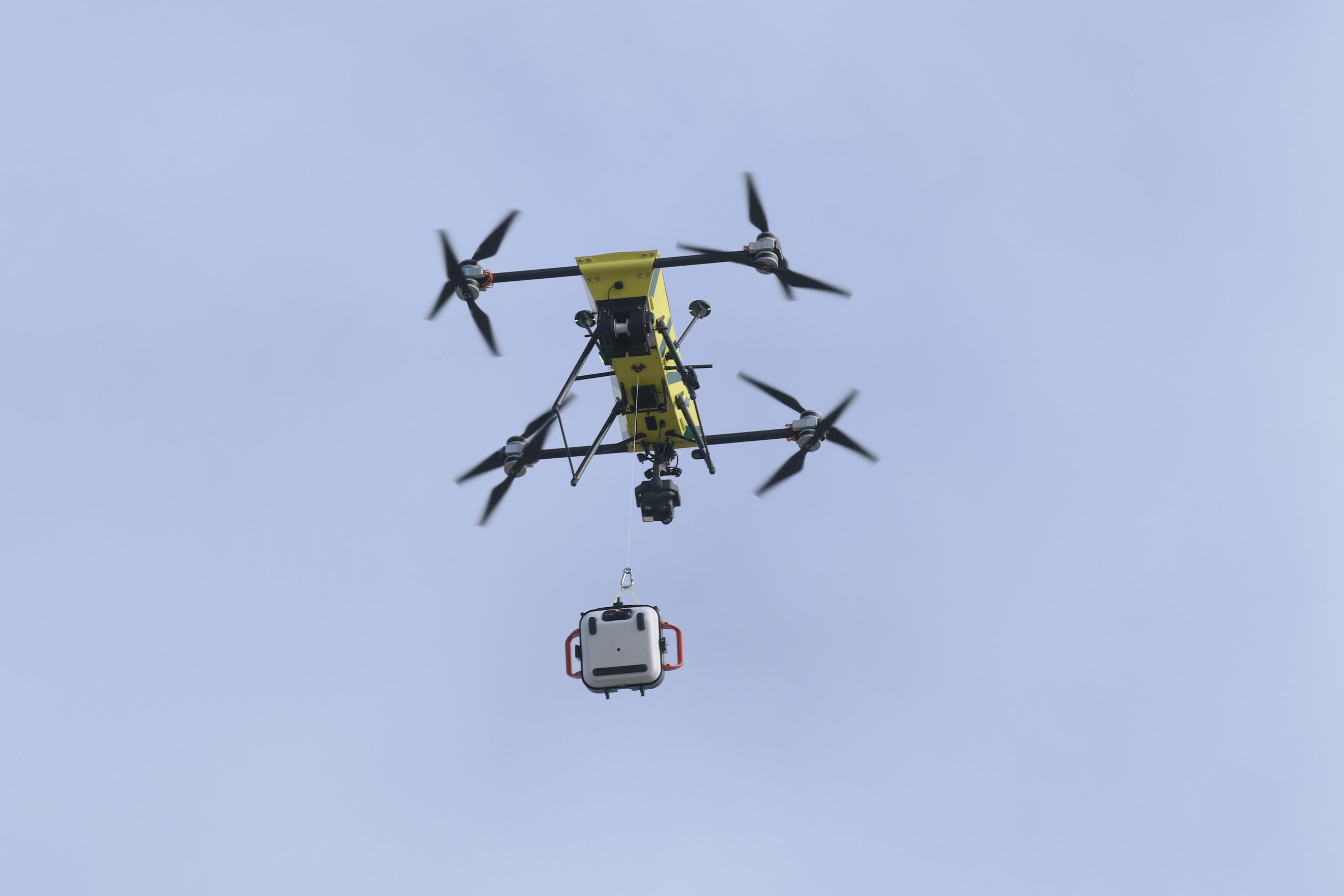 Yellow multi-rotor drone carrying a white container suspended beneath it against a clear blue sky.