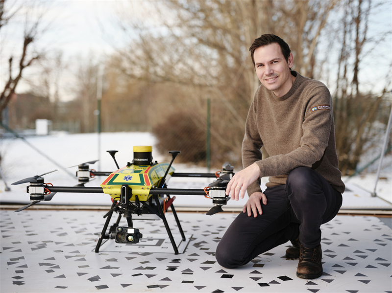 Man kneels beside a large yellow quadcopter drone on a checkered platform outdoors in winter.