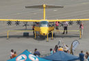 Yellow small twin-engine airplane on a tarmac at an outdoor airshow, with people inspecting it behind yellow barriers.