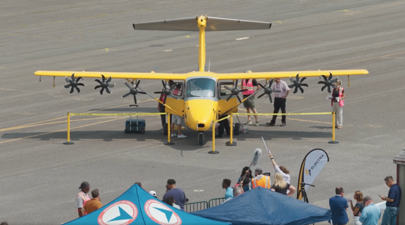 Yellow small twin-engine airplane on a tarmac at an outdoor airshow, with people inspecting it behind yellow barriers.