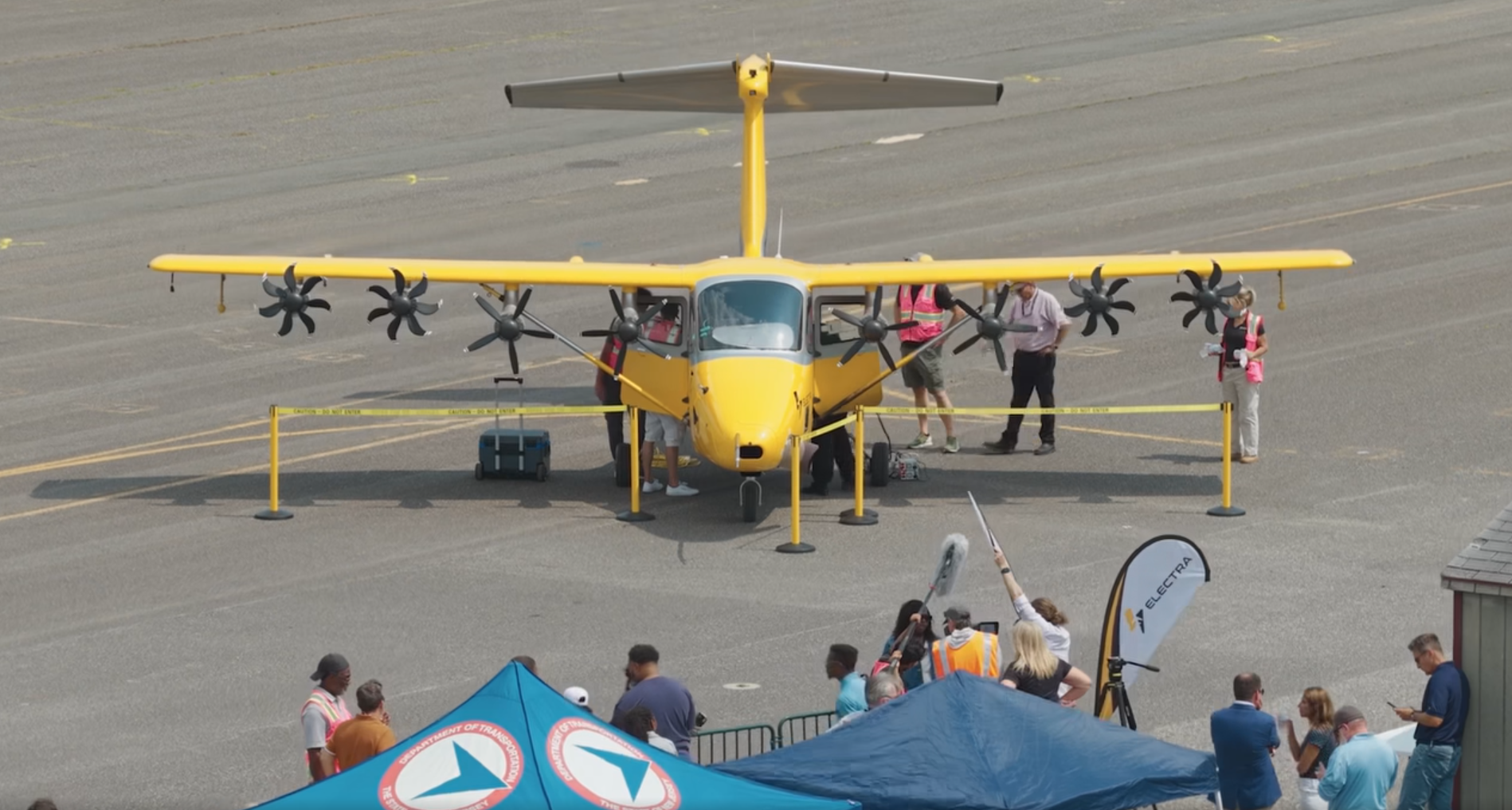 Yellow small twin-engine airplane on a tarmac at an outdoor airshow, with people inspecting it behind yellow barriers.