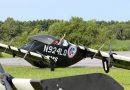 Pilot in a black biplane on a runway giving a thumbs-up, aircraft labeled N924LQ with an emergency services patch visible on the fuselage.