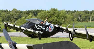 Pilot in a black biplane on a runway giving a thumbs-up, aircraft labeled N924LQ with an emergency services patch visible on the fuselage.